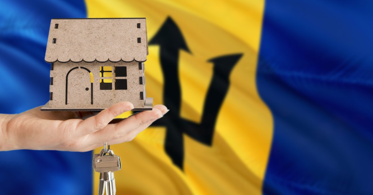 A person holding a small wooden house and keys in front of the Barbados flag as it billows in the background.