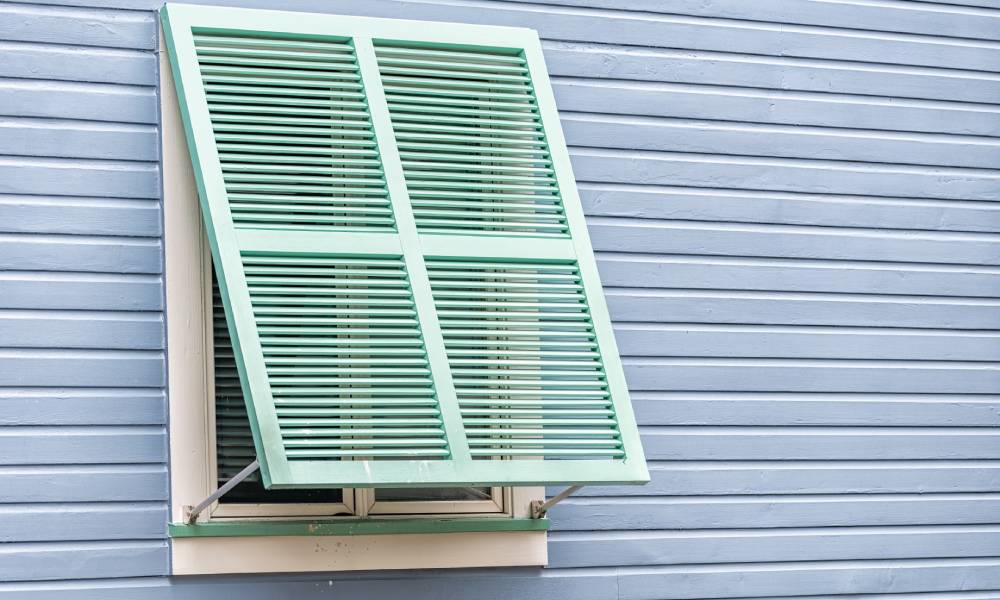 Turquoise storm shutters that are slightly ajar on the window of a light blue house in the Caribbean.